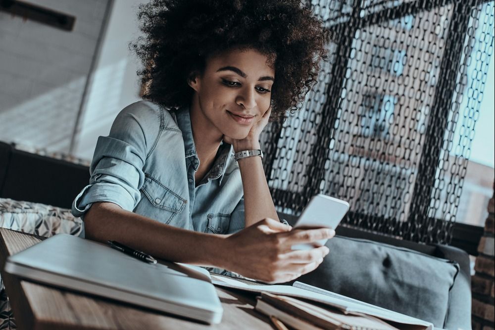 african-woman-looking-at-smart-phone-and-smiling-while-sitting-in-in-cafe-or-creative-office